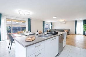 A modern kitchen with a central island, a dining area with a table and chairs, and a living room with a sofa and a TV. - Spacious 4B House in PointCook (Point Cook)