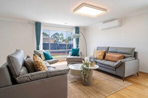 A modern living room with a grey sofa set, a coffee table, and a large window with curtains. - Spacious 4B House in PointCook (Point Cook)