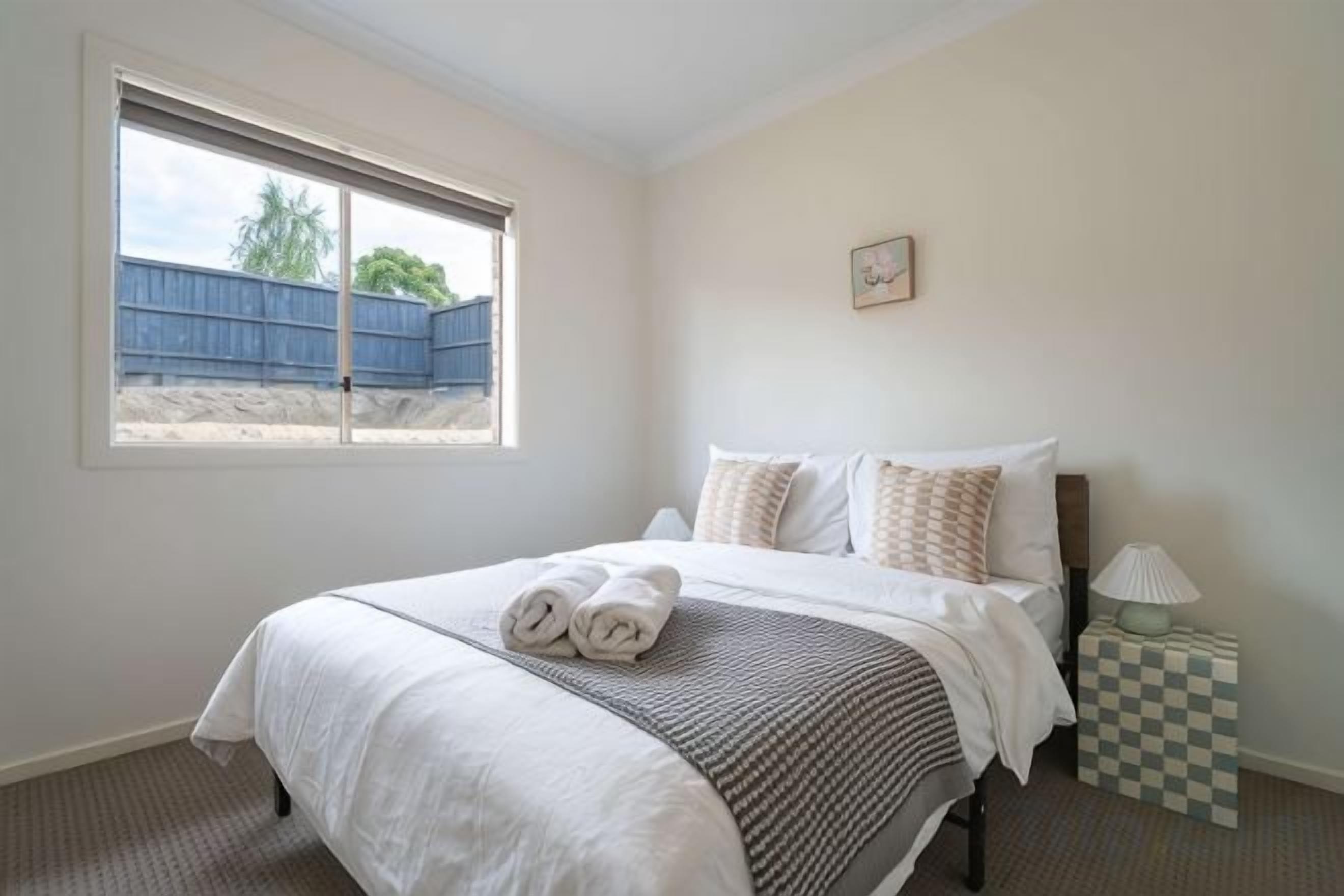 Un dormitorio con cama, almohadas, una mesita de noche con lámpara, y ventana con vista a una valla azul y árboles.