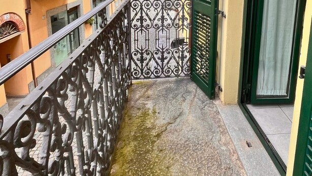 Un balcón estrecho con barandilla de metal, una puerta con contraventanas verdes y vistas a edificios vecinos.