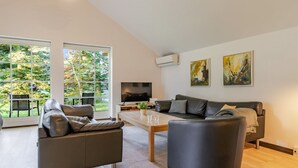 Ett modernt vardagsrum med en soffa, fåtölj och soffbord, stora fönster med utsikt över grönområden och två abstrakta tavlor på väggen.