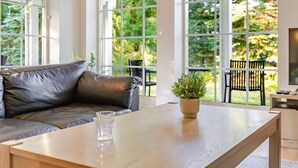 Ett modernt vardagsrum med en soffa i läder, ett soffbord i trä och stora fönster som ger utsikt över grönområden utanför.