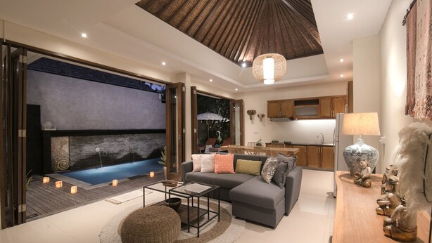 A modern living room with a grey sofa, a wooden coffee table, and a round pouf. There is a kitchen area with wooden cabinets and a dining table. A large window provides a view of the outdoors. - Marijo Villa 2BR Private Pool Mins to Central Ubud (Ubud)