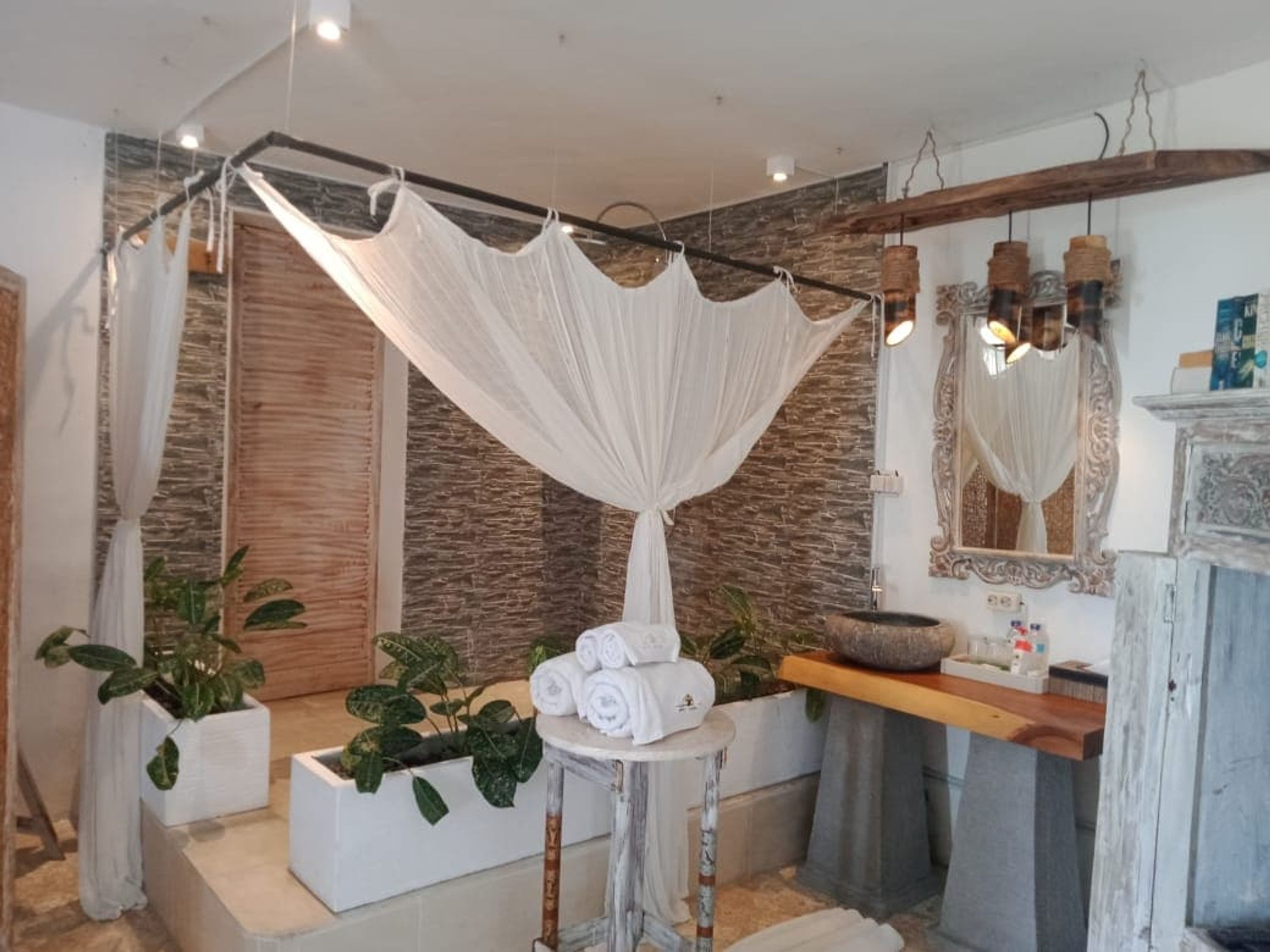 Une salle de bain avec un mur en pierre, un ciel de lit blanc, un meuble-lavabo en bois et des plantes en pot.