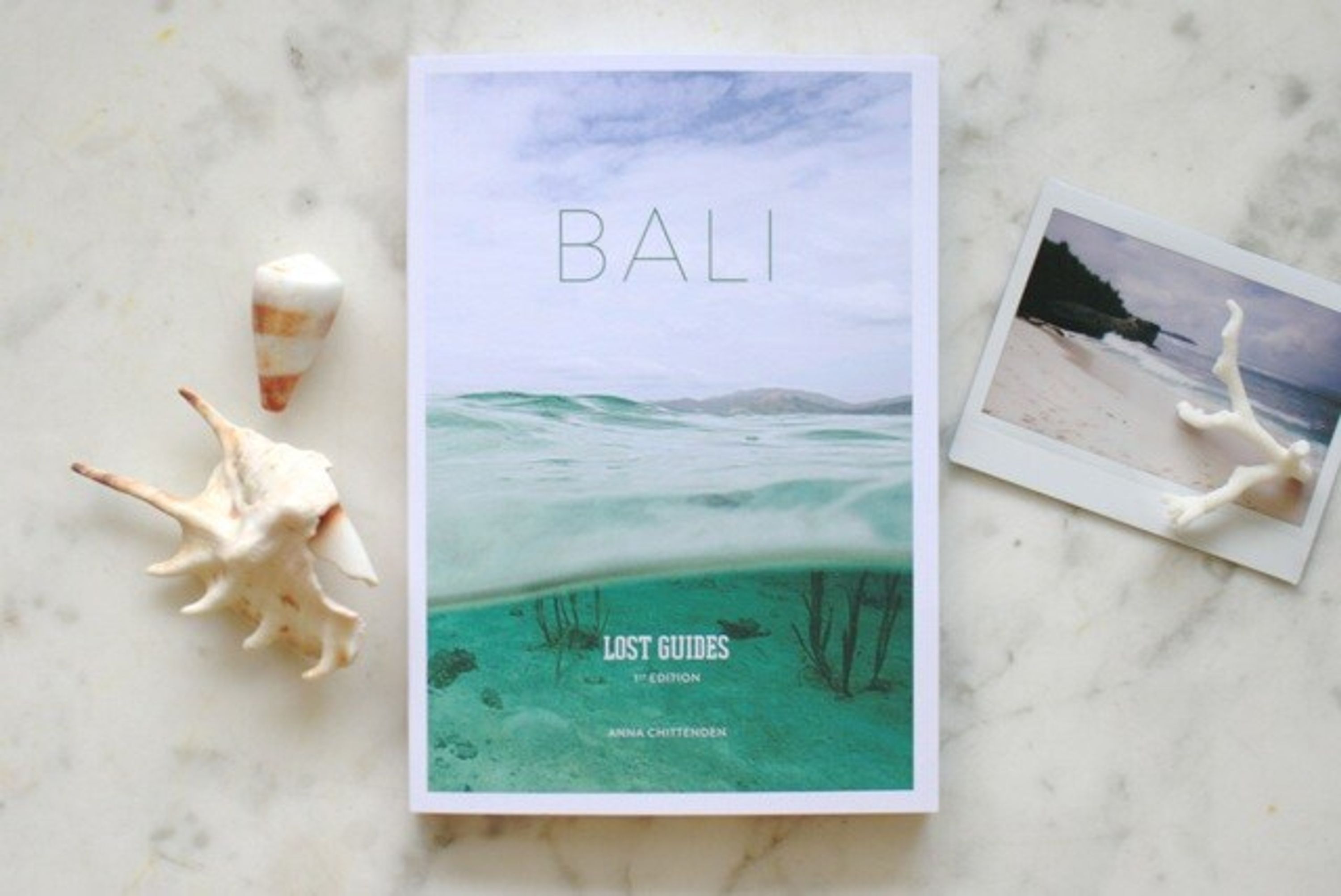 Un libro titulado 'BALI LOST GUIDES' de Anna Chittenden sobre una superficie de mármol con caracolas y una fotografía tipo Polaroid.
