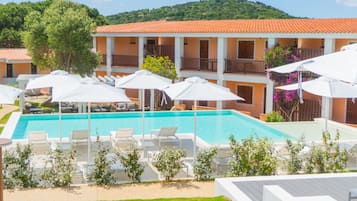 Un área de piscina con sillones de descanso y sombrillas, un edificio con balcones y una ladera verde al fondo.
