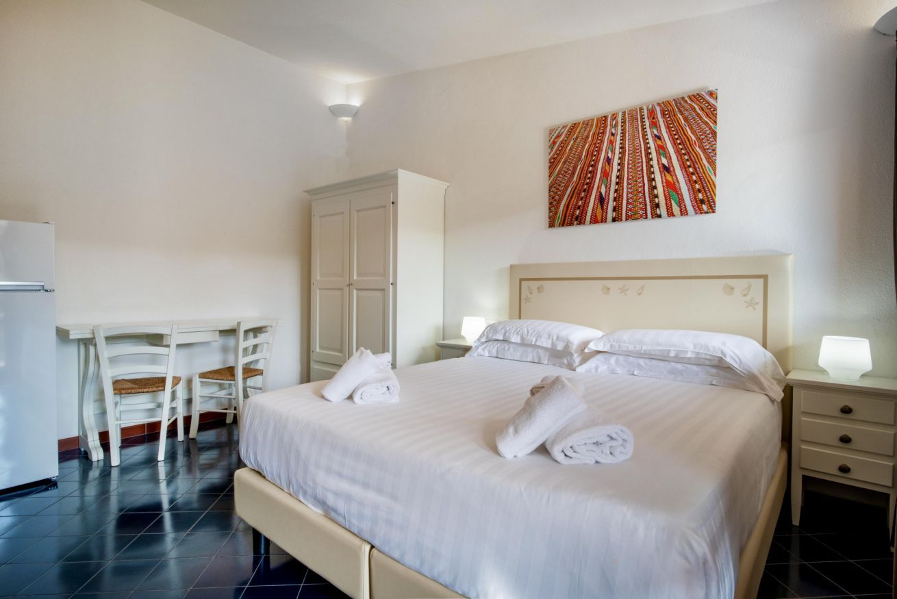 A hotel room with a bed, white curtains, a desk with chairs, a wardrobe, and a wall-mounted artwork.