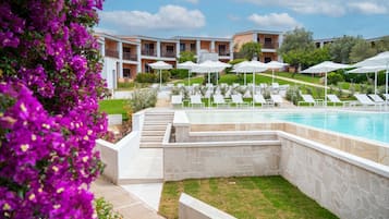 Zona de piscina con sillones de descanso y sombrillas, rodeada por un muro de piedra y un edificio con balcones.