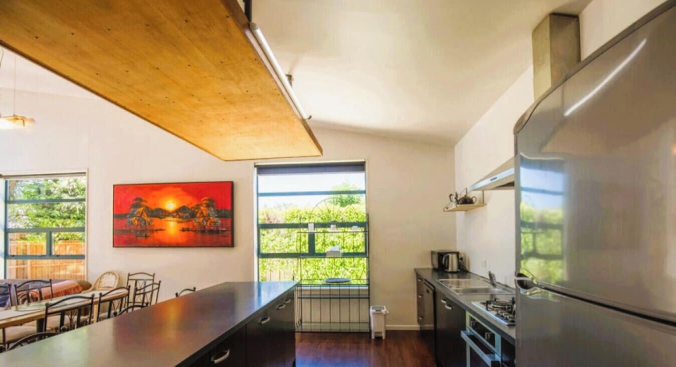 Uma cozinha moderna com uma geladeira grande, uma área de refeições com uma mesa comprida e cadeiras, e vista para o verde através de grandes janelas.