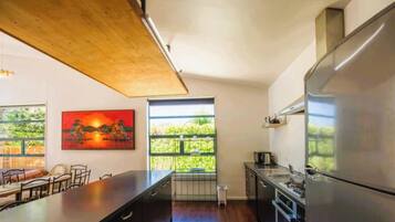 Uma cozinha moderna com uma geladeira grande, uma área de refeições com uma mesa comprida e cadeiras, e vista para o verde através de grandes janelas.