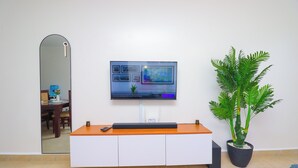 A modern living room with a flat-screen TV mounted on a white wall, a wooden coffee table, a potted plant, and a dining area visible through a glass door.