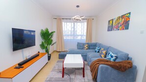 Ein modernes Wohnzimmer mit Flachbildfernseher, einem Holztisch, einem blauen Sofa mit gemusterten Kissen, einem weißen Couchtisch und einem großen Fenster mit Vorhängen.