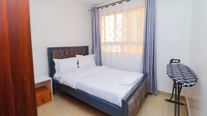 A neatly made bed with white linens, a wooden nightstand, a small wooden cabinet, and a black ironing board with a patterned cover.