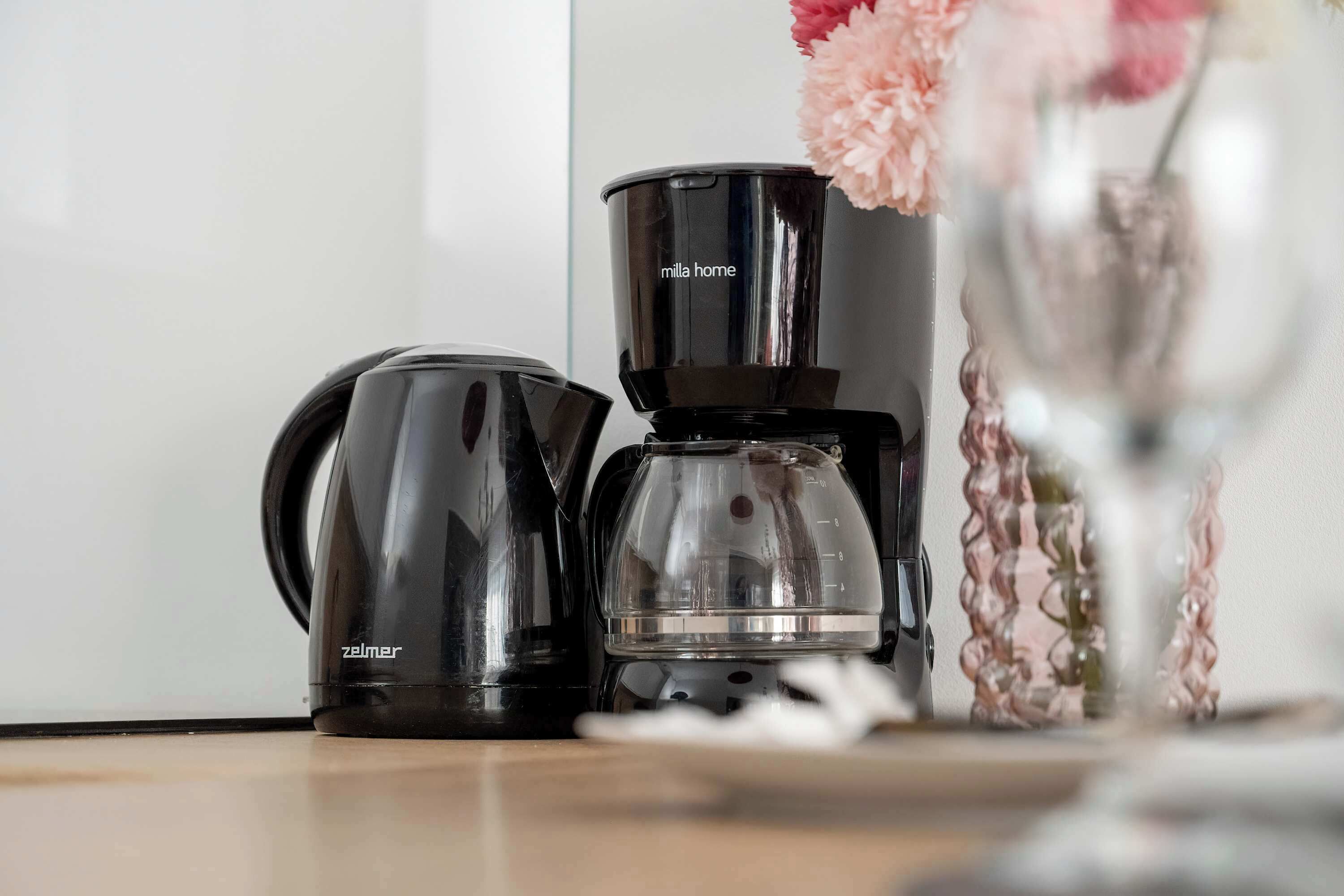 A coffee maker and a kettle on a table with a vase of flowers in the background.