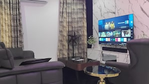 A modern living room with a flat-screen TV, a sofa, a coffee table, and a fan. - Dwellux Apartments  (Abuja)