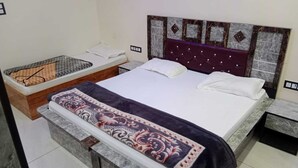 A bedroom with a bed, a bedside table, a nightstand, and a small seating area with a cushion. - Hotel Radhe Krishna (Khatoo)
