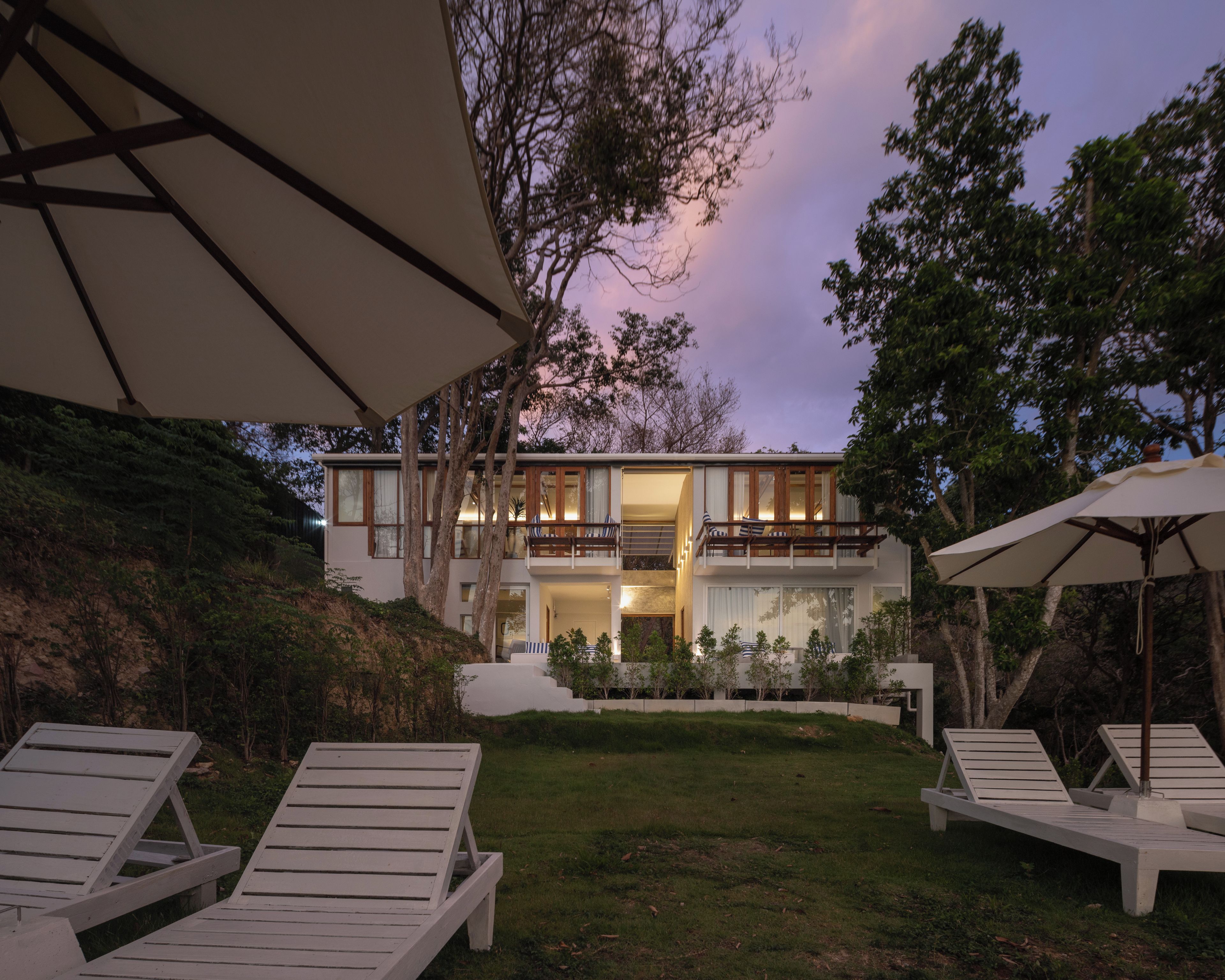 A modern two-story house with a balcony, a lawn, and lounge chairs.
