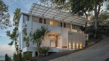 A modern two-story building with a flat roof, large windows, and a balcony. There are potted plants and a pathway leading to the entrance.