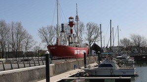 Ein großes, rotes Schiff liegt in einer Marina vor Anker, im Hintergrund ist ein Leuchtturm zu sehen.
