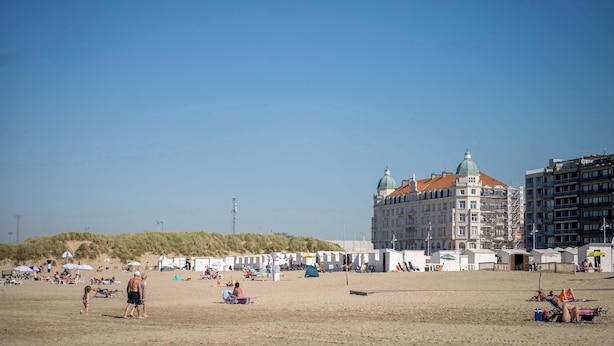 Ein Strand mit Zelten und Sonnenschirmen, ein großes Gebäude mit Kuppel und eine Sandfläche mit Menschen.