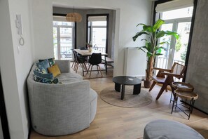 A spacious living area with a grey sofa, a dining table, and a large potted plant. - Belle Époque Apartment - Steps to Blankenberge (Blankenberge)