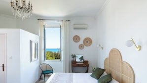 Ein Schlafzimmer mit Bett, Sessel, Schreibtisch, einer Chandelier und Blick auf das Meer.