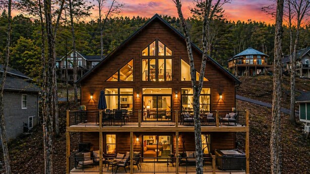 Una cabaña de madera de dos pisos, con amplias ventanas, un balcón y una terraza con mobiliario de exterior.