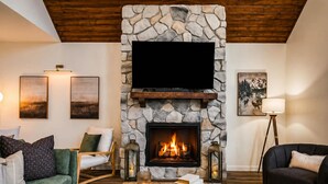 Una sala de estar acogedora con una chimenea de piedra, un televisor de pantalla plana montado encima y asientos cómodos, incluyendo un sofá verde y un sofá oscuro.