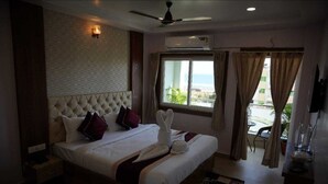 A hotel room with a large bed, a ceiling fan, an air conditioning unit, a window with curtains, and a view of the outdoors. - New Beach Resort (Brahmagiri)
