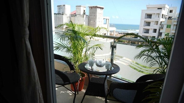 A balcony with a table and chairs, a potted plant, and a view of the ocean and buildings. - New Beach Resort (Brahmagiri)