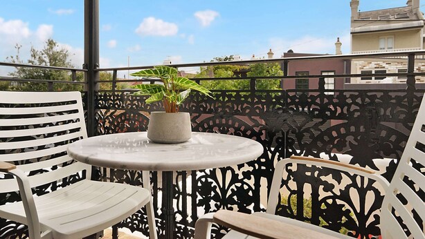 A balcony with a round table and two white chairs, a potted plant, and a decorative metal fence. - Cozy Studio With Balcony in Unbeatable Location (Sydney)