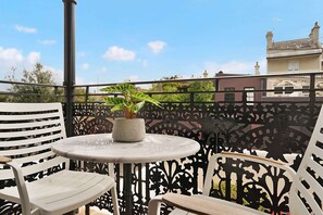 Un balcone con un tavolo rotondo e due sedie bianche, una pianta in vaso e un recinto metallico decorativo.