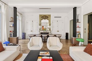 Een ruime woonkamer met een open haard, twee witte fauteuils, een salontafel met een tijdschrift, en een eethoek met een kroonluchter.