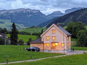 A two-story wooden house with a sloped roof, a visible chimney, and a car parked in front. The house is surrounded by a green landscape with mountains in the background. - Panoramic Views & Appenzell Charm: Your Spacious Retreat (Appenzell)