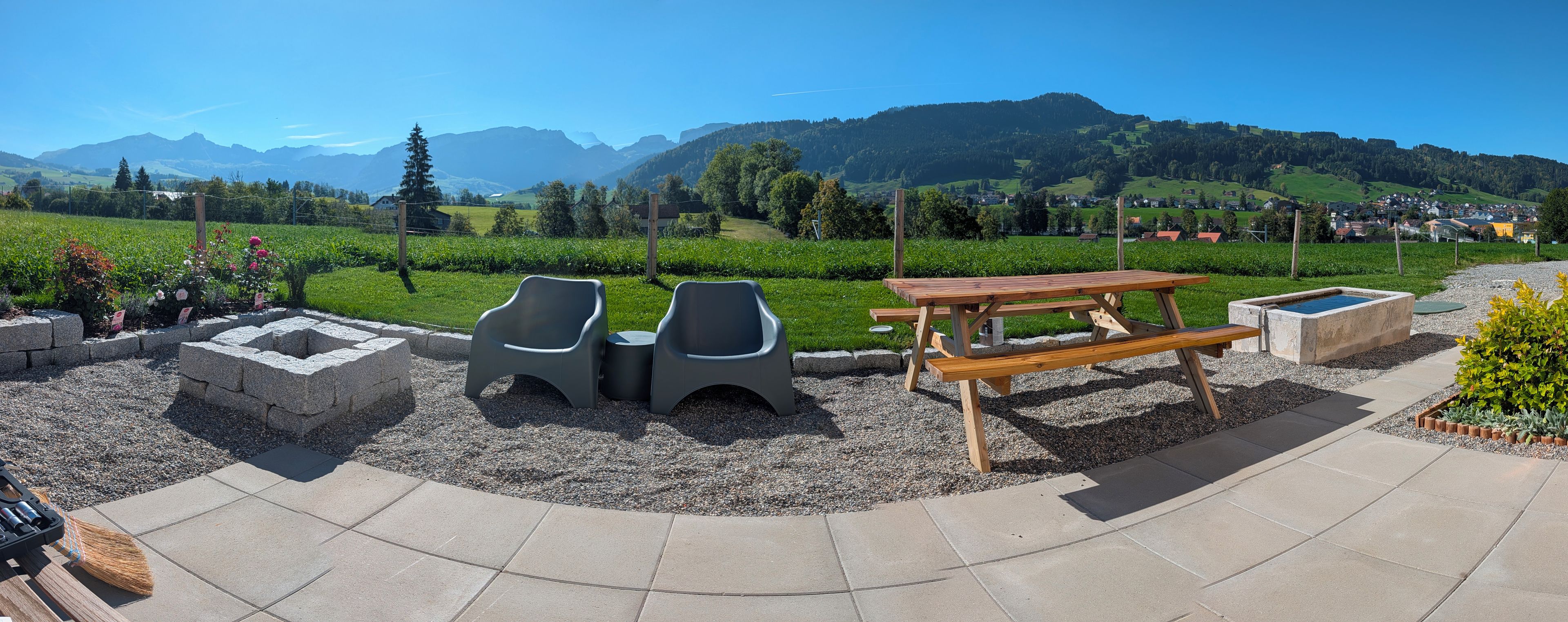 An outdoor seating area with a picnic table and two chairs, surrounded by a gravel path and a grassy field with a mountain backdrop.
