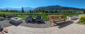 An outdoor seating area with a picnic table and two chairs, surrounded by a gravel path and a grassy field with a mountain backdrop.