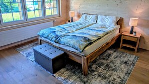 A bedroom with a wooden bed, two bedside lamps, a window with a view of greenery, and a small bench.