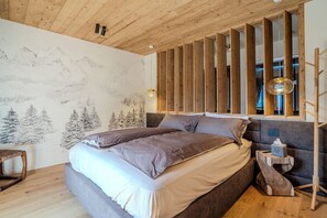 A bedroom with a bed, wooden headboard, a wall mural of mountains, a bedside table with a lamp, and a small wooden stool. - Roccia Nera Luxuty Suite Champoluc Monterosa Ski (Ayas)