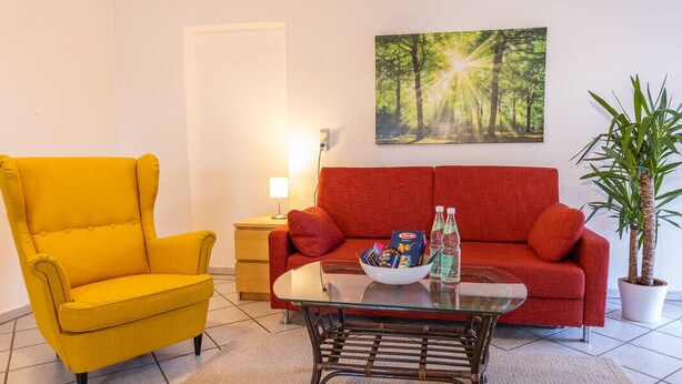 Un soggiorno con una poltrona gialla, un divano rosso, un tavolino da caffè in vetro e un quadro raffigurante una foresta.
