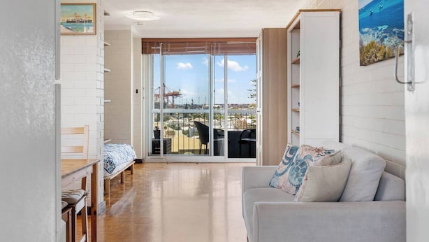 A modern living room with a sofa, a dining table, and a view of the city through large glass doors. - River Vistas - Fremantle Port Views (North Fremantle)