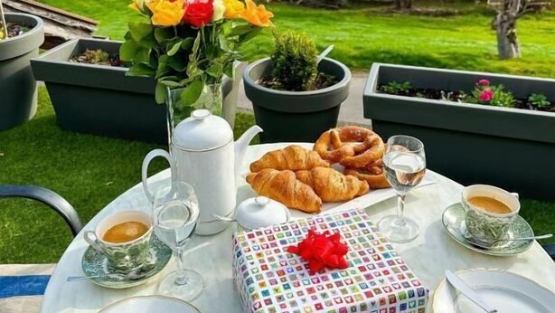 Mesa para comer al aire libre con croissants, tetera y una caja de regalo, con un paisaje de montañas al fondo.