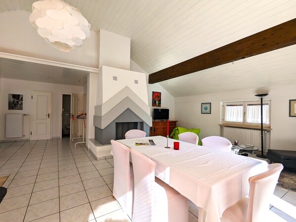 Un comedor con una mesa blanca dispuesta para cuatro personas, chimenea y techo alto con vigas de madera a la vista.