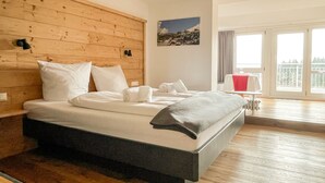Una habitación de hotel moderna con cabecera de madera, una cama grande con sábanas blancas, una pequeña mesa con mantel rojo y un balcón con vistas a árboles.