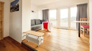 Un salón moderno con un sofá gris, una pequeña mesa con mantel rojo y una silla de madera. Hay un radiador debajo de un banco de madera y una ventana grande con cortinas.