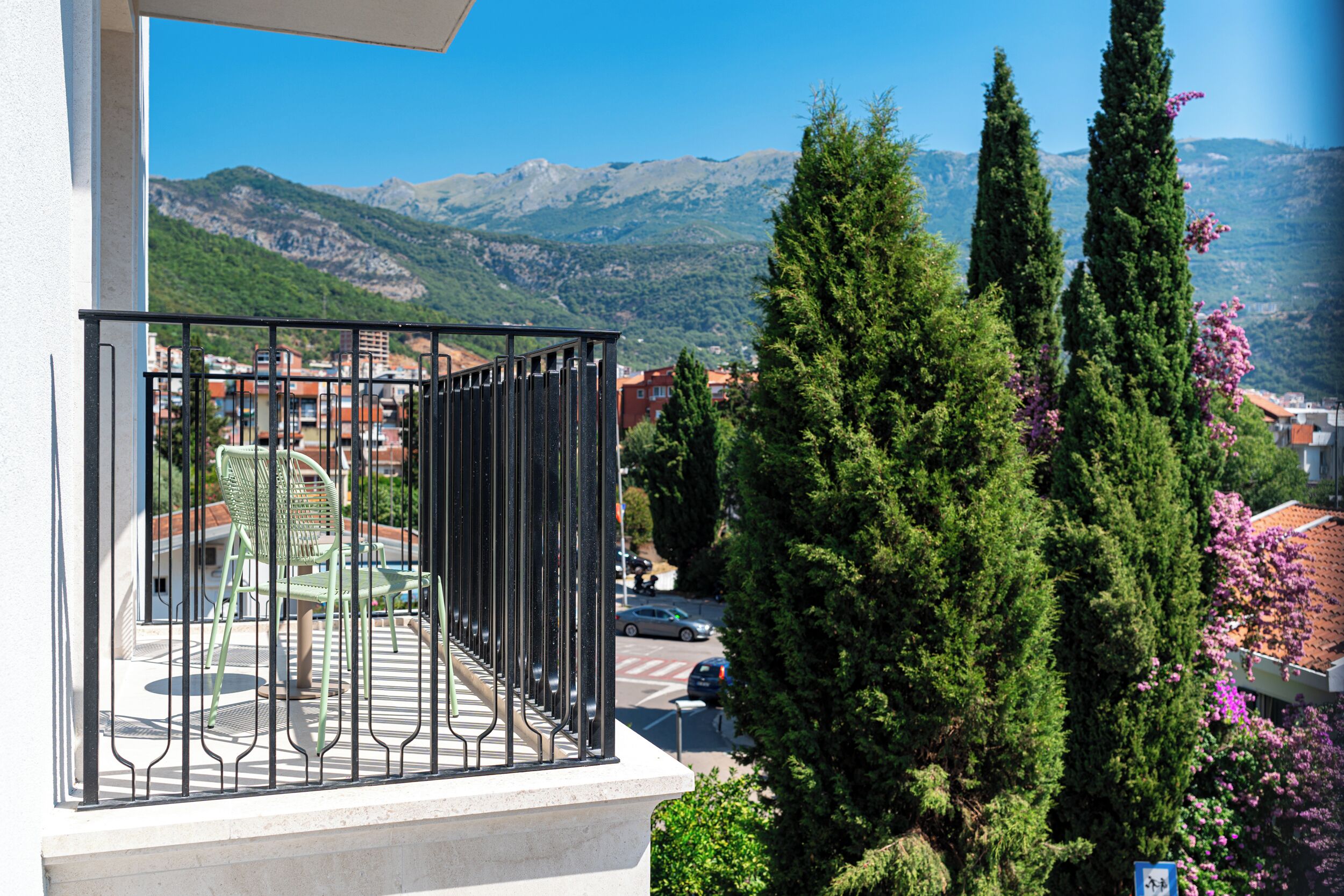 A balcony with a metal railing, a table, and chairs, overlooking a street with cars and buildings, with mountains in the background.