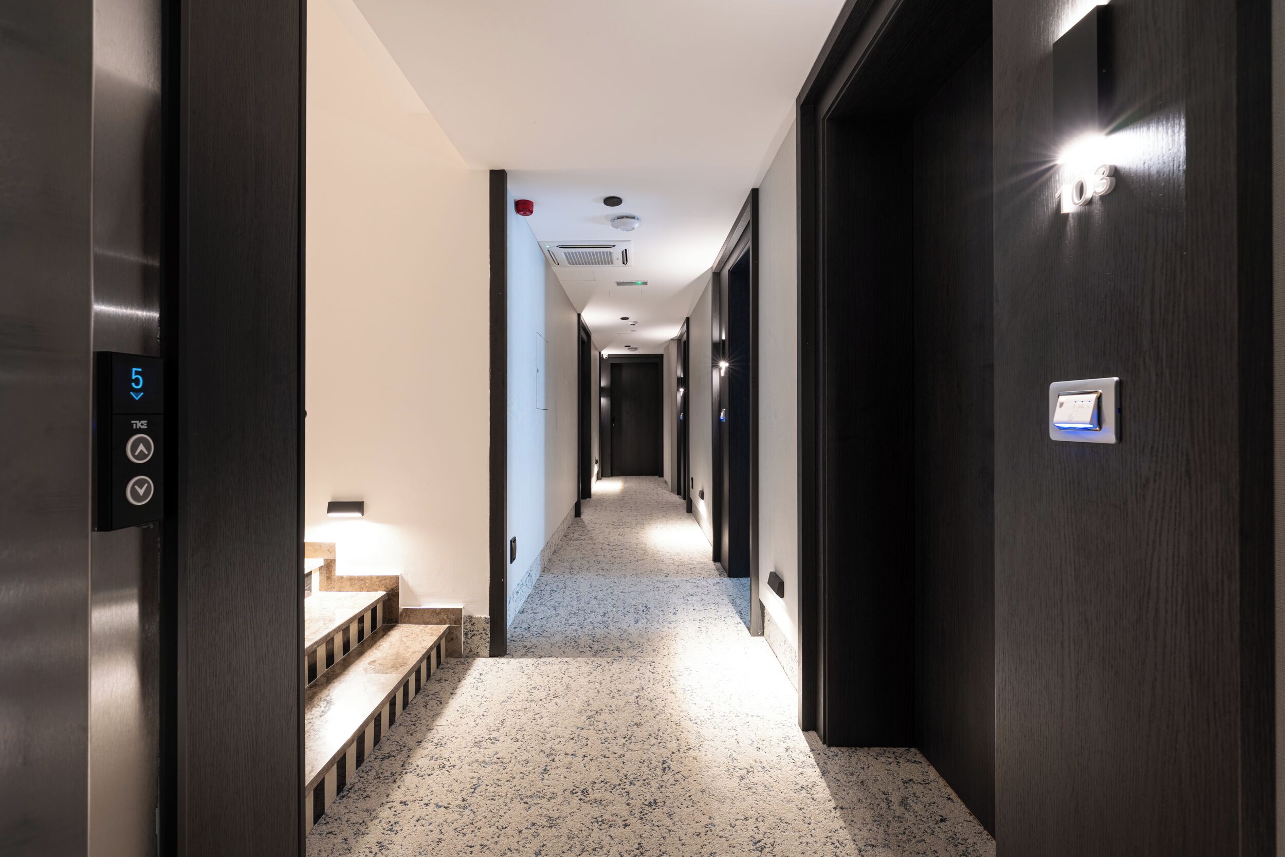 Un pasillo de hotel de diseño moderno, con una escalera, un panel digital de ascensor y un largo corredor con puertas a un lado.