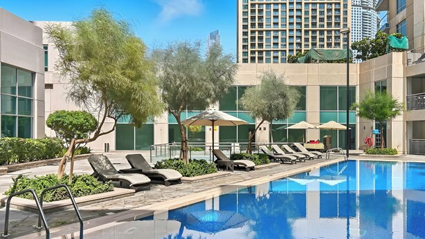 A swimming pool with lounge chairs and umbrellas, surrounded by trees and modern buildings. - Studio Near Dubai Mall Burj Khalifa (Dubai)