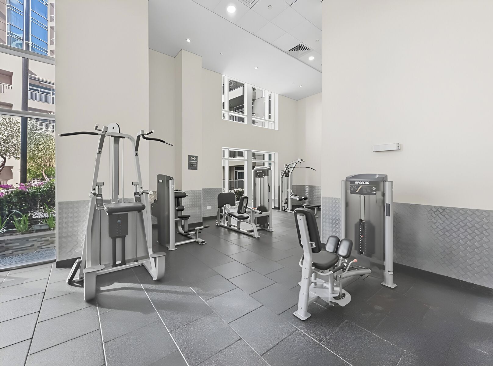 Un gimnasio con máquinas de ejercicio variadas, un espejo grande y vistas a un edificio exterior.