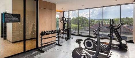 Uma academia com equipamentos de ginástica, um suporte com halteres e uma janela grande com vista para a cidade.