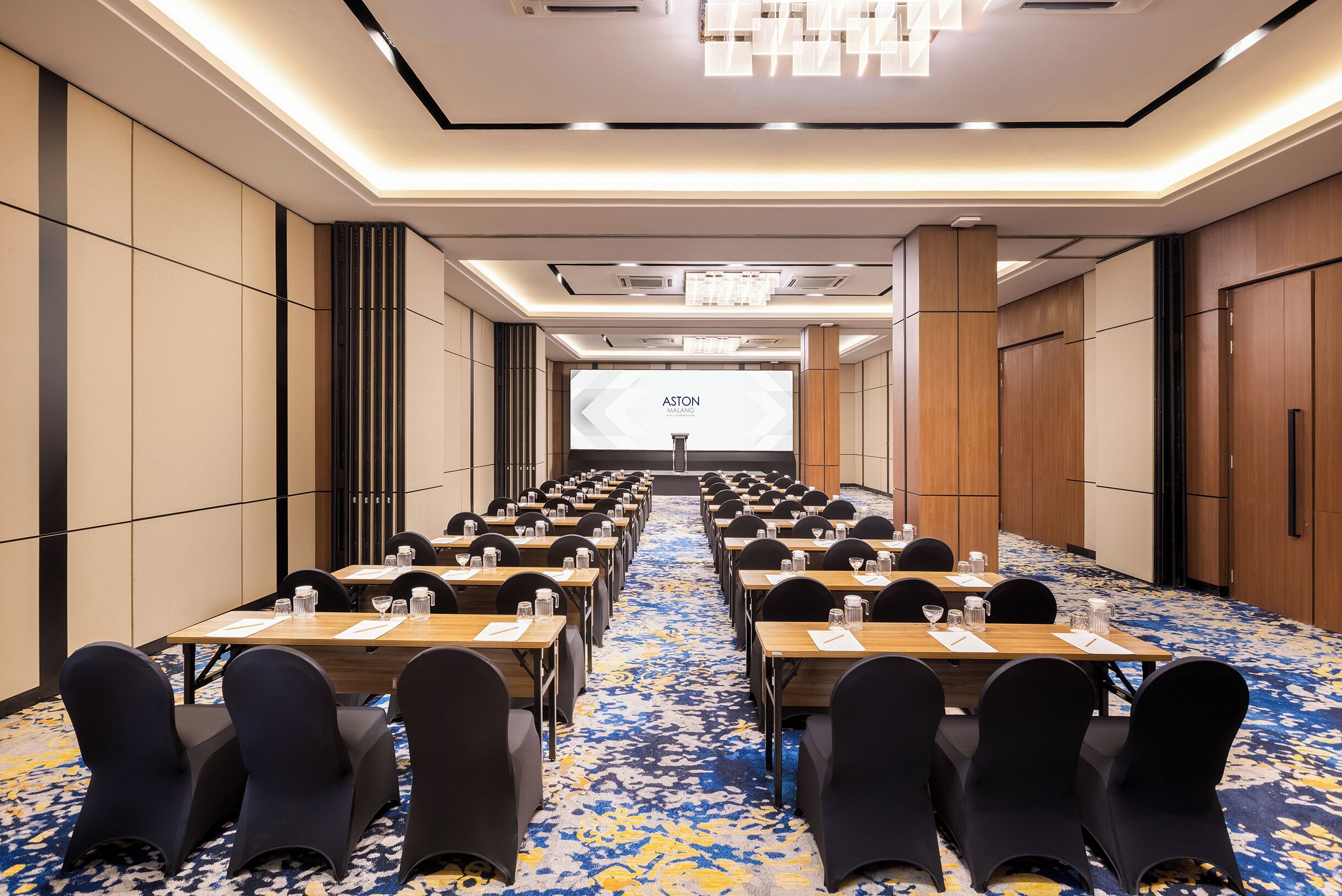 A conference room with rows of tables and chairs, a projector screen, and a carpeted floor.